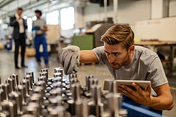 Young steel worker examining manufactured rod cylinder while using digital tablet in distribution warehouse